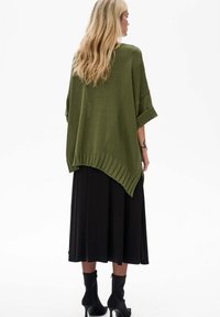 Zielona oliwkowa oversize'owa bluzka dzianinowa z prążkowanym dołem, zestawiona z czarną plisowaną spódnicą. Posiada luźne rękawy i fakturowany materiał.