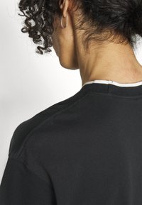 Svart bomullströja med rund halsringning, vit sömdekoration, slät yta och förstärkta sömmar vid axlarna.