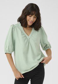 Blouse vert clair avec un décolleté en V, des manches bouffantes et un tissu texturé. Coupe décontractée avec des détails froncés au niveau du col et des manches.