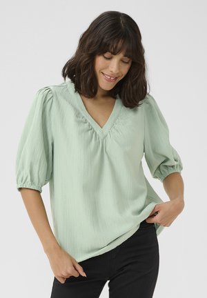 Blouse vert clair avec un décolleté en V, des manches bouffantes et un tissu texturé. Coupe décontractée avec des détails froncés au niveau du col et des manches.