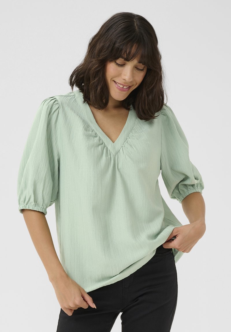 Blouse vert clair avec un décolleté en V, des manches bouffantes et un tissu texturé. Coupe décontractée avec des détails froncés au niveau du col et des manches.