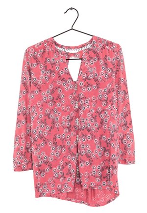 Blouse rose à manches longues avec des motifs floraux circulaires blancs et noirs, boutonnée sur le devant, avec un petit décolleté en forme de serrure, suspendue sur un cintre noir.