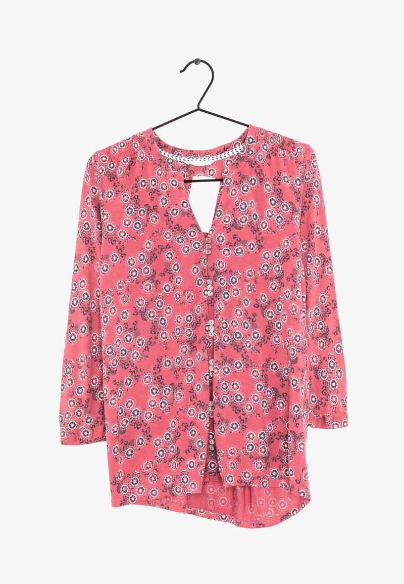 Blouse rose à manches longues avec des motifs floraux circulaires blancs et noirs, boutonnée sur le devant, avec un petit décolleté en forme de serrure, suspendue sur un cintre noir.