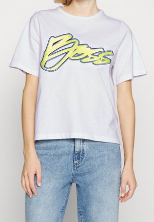 Osoba w jasnofioletowej koszulce z żółtym i niebieskim napisem „Boss” oraz jasnoniebieskich dżinsach, ręce swobodnie opuszczone przy bokach.