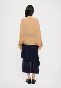 Nainen, jolla on pitkät tummat hiukset, seisoo selin päälläsi läpinäkyvä beige viitta ja kerroksellinen tummansininen hame mustilla kengillä.