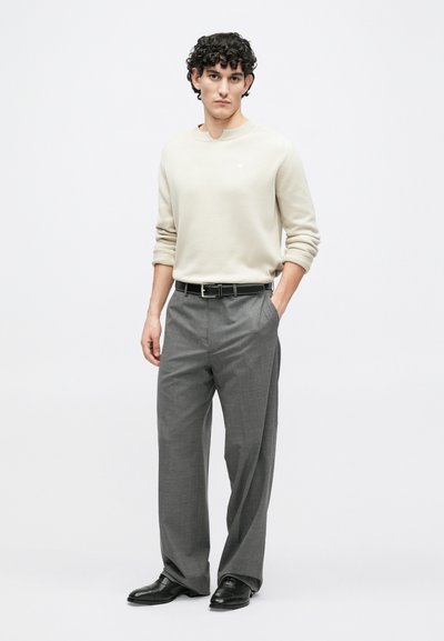 Ελαφρύ μπεζ πλεκτό πουλόβερ με στρογγυλή λαιμόκοψη, γκρι παντελόνια με σχέδια, μαύρη δερμάτινη ζώνη και παπούτσια, φορεμένα από ένα πρόσωπο που στέκεται μπροστά σε απλό φόντο.