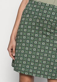 Zelená sukně s vzorem obsahuje květinové motivy v červené a bílé barvě na černém pozadí. Materiál se zdá být lehký a texturovaný, s rozšířeným tvarem.