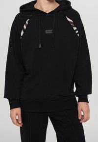 Černá mikina s kapucí s žebrovanými manžetami a lemem, s výřezy na bocích odhalujícími proužkovaný vzor v hnědé a krémové barvě. Na hrudi je nášivka s logem.