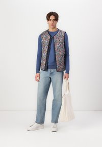 Floral patterned padded vest with navy trim, worn over a blue long-sleeve top, paired with light blue jeans and white sneakers, holding a tote bag.