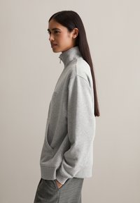 Grauer Reißverschluss-Sweatshirt mit hohem Kragen, featuring eine Fronttasche, gerippte Bündchen und eine lockere Passform, getragen über karierten Hosen.