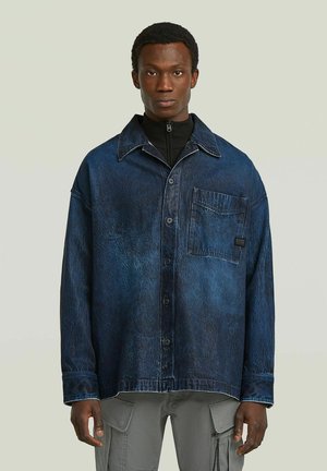 Junger Mann, der eine dunkelblaue Jeansjacke mit einer Vordertasche über einem schwarzen Reißverschluss-Pullover und grauen Cargohosen trägt, vor einfachem Hintergrund.