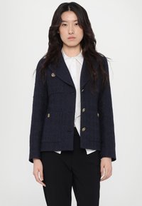 Granatowa tweedowa kurtka z złotymi guzikami, krótki krój, kieszenie naszyte i pleciona faktura. Nosi się ją na białej koszuli.