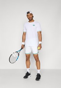 Valge tennisekomplekt roheliste detaljadega, sisaldab T-särki ja lühikesi pükse. Komplektil on mustad tenniseketsid ja reket kollase palliga.