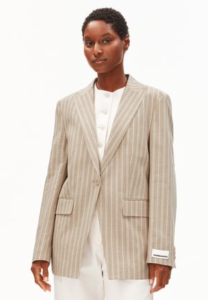 Vrouw draagt een beige krijtstreepblazer over een wit overhemd met knopen, staand tegen een effen witte achtergrond.