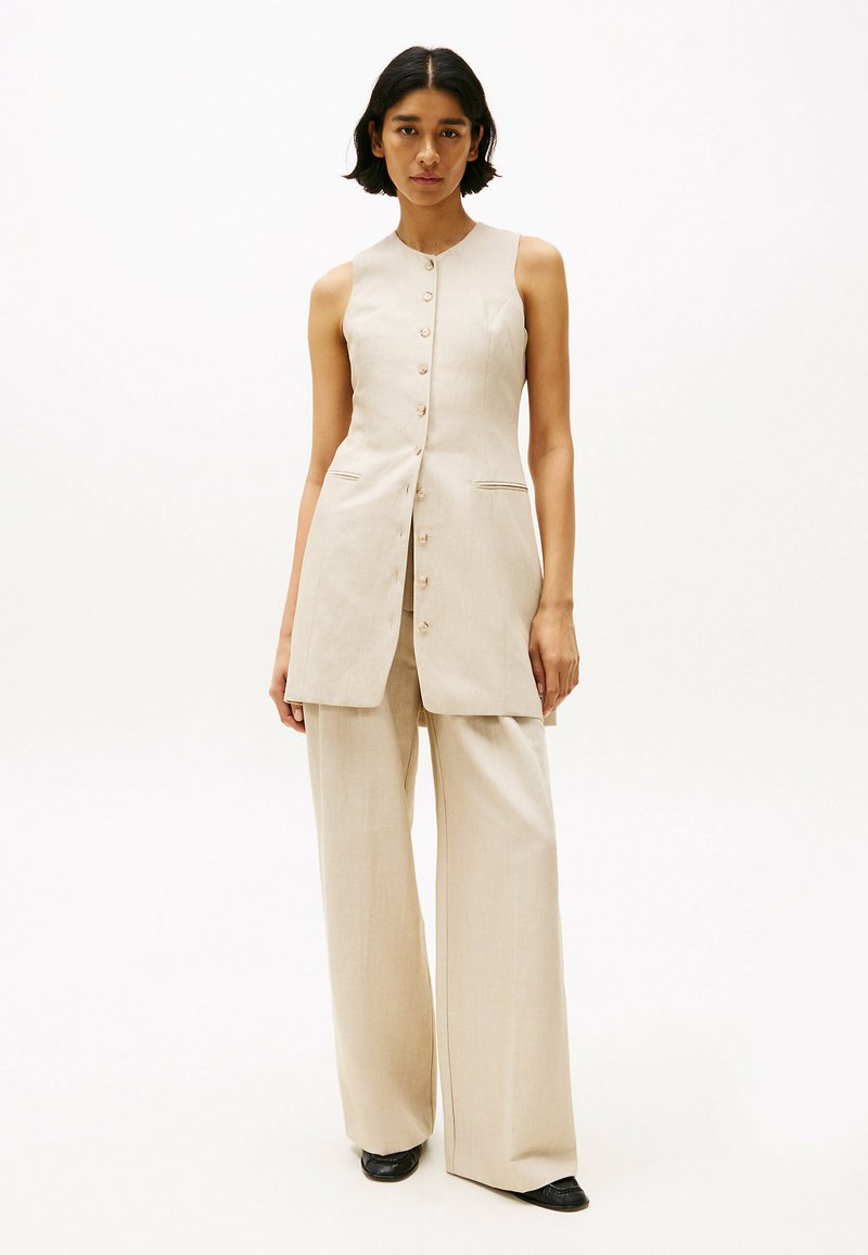Tunique sans manches beige clair à coupe droite, avec fermeture à boutons et deux poches latérales, associée à un pantalon large beige. Chaussures noires.