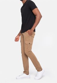 Beige cargobroek met meerdere zakken, taps toelopende pasvorm en een gladde textuur, gecombineerd met een nauwsluitend zwart T-shirt en witte sneakers.