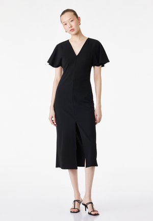 Ung kvinde med kort hår iført en sort midi-kjole med flagrende ærmer, frontslids og sorte sandaler med hæl og stropper.