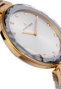 Reloj en tono dorado que presenta un bisel de cristal gris facetado, una esfera plateada con marcadores de diamantes y manecillas doradas, con una pulsera de eslabones metálicos.