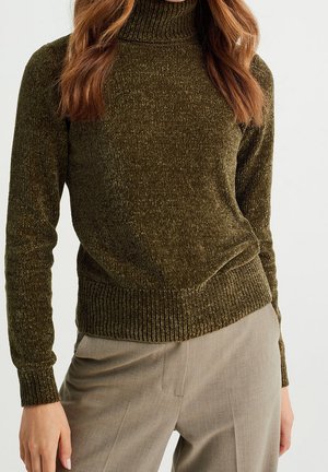 Femme portant un pull à col roulé en chenille verte avec poignets et ourlet côtelés, associé à un pantalon beige sur mesure sur un fond uni.