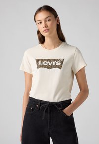 Kreminės spalvos marškinėliai su leopardų rašto Levi's logotipu. Trumpomis rankovėmis ir klasikiniu apvaliu kaklu. Suderinti su juodomis, aukštu liemeniu kelnėmis.