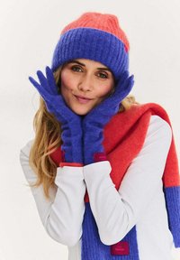 Čepice z pleteniny se růžovými a modrými pruhy; fialové rukavice a červená šála s odpovídající cedulkou. Měkké, texturované materiály vytvářejí útulný vzhled.