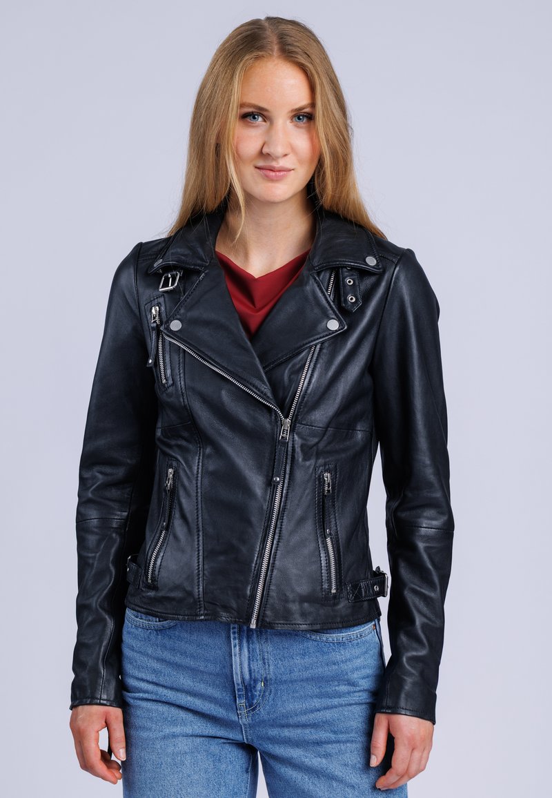 Ung kvinde med langt blond hår iført en sort læderjakke over en rød top og blå jeans, stående mod en ensfarvet baggrund.