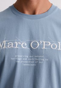 Blauwe katoenen t-shirt met witte geborduurde tekst. Het ontwerp bevat de merknaam "Marc O'Polo" en een boodschap over natuurerfgoed.