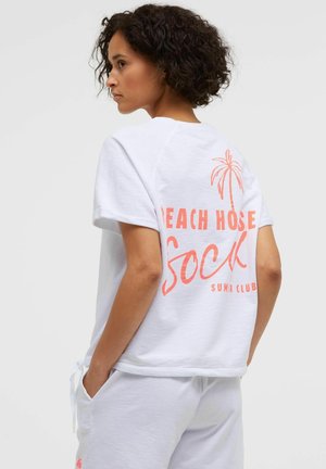 Frau mit kurzen lockigen Haaren, die ein weißes T-Shirt mit korallenfarbenem Text und Palmenmotiv sowie weiße Hosen trägt, mit den Händen in den Taschen, seitlich stehend.