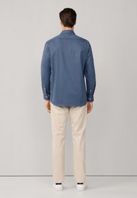 Heren blauwe button-up overhemd in textiel met textuur, lange mouwen, achteraanzicht. Gepaarde met beige broeken en witte schoenen. Eenvoudig ontwerp met een kraag.