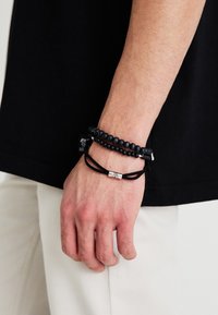 Melnās pērļu rokassprādzes uz plaukstas, kurām ir gludas un teksturētas pērlītes, ar sudraba akcentu uz melna auklas. Roka daļēji redzama.