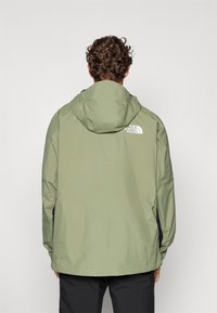 Helle grüne Regenjacke für Herren mit Kapuze, ausgestattet mit einem weißen Logo auf dem oberen Rücken, aus einem glatten, wasserabweisenden Material, minimalistisches Design.
