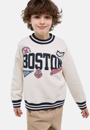 Garçon souriant aux cheveux bouclés, portant un sweat-shirt blanc avec le texte "BOSTON" et des écussons à thème nautique et sportif, pantalon beige, debout les bras légèrement écartés.