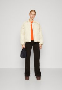 Puffy kremjakke, tettsittende oransje topp, svarte bukser og brune tresko. Modellen holder en mørkeblå teksturert håndveske.