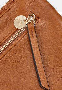 Brun læder håndledtaske med guld lynlås og rund guldlogo-charms, der er fastgjort til lynlåsens trækflap.