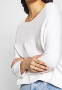 Femme portant un sweat à manches longues blanc et un jean bleu, bras croisés à la taille, sur un fond clair uni.