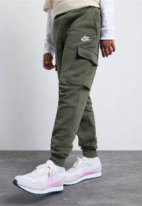 Person som bär olivgröna Nike cargo joggers och vita sneakers med rosa Nike swooshar på ett grått golv och vit bakgrund.