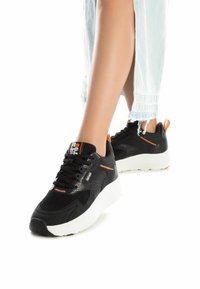 Zapatillas deportivas negras con malla transpirable y acentos de cuero, con una suela blanca gruesa y detalles en naranja en el talón y la lengua.