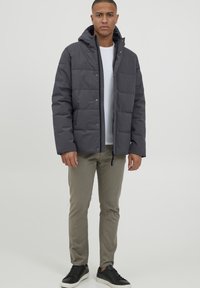 Grå pufferjakke med hætte, der har et quiltet design og trykknaplukninger. Sammen med lyse bukser og sorte sneakers.