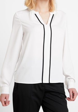 Witte blouse met lange mouwen en zwart verticale randdetails aan de voorkant, gecombineerd met zwarte broek, gedragen door een staand persoon.