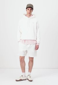 Sudadera blanca con cordones sobre una camisa rosa con cuello, combinada con pantalones cortos blancos texturizados y zapatillas beige. Una gorra a cuadros completa el look.