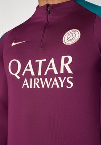 Violetti urheilu-paita, jossa on vetoketjokaulus, valkoisella "QATAR AIRWAYS" -tekstillä, logo-patchi ja teal-värit olkapäissä.