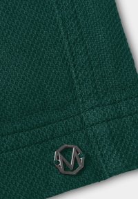 Donkergroen gebreide stof met structuur en een klein metalen achthoekig logo met de letter "M" dicht bij de rand genaaid.