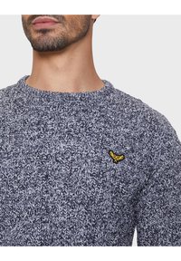 Pletený svetr v tmavém melírovaném vzoru s kulatým výstřihem a malým žlutým emblémem na hrudi. Texturovaný materiál s útulným vzhledem.