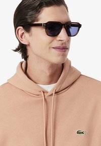 Felpa marrone con cappuccio e coulisse, con un piccolo logo di coccodrillo verde sul petto; occhiali da sole rettangolari con lenti scure e montatura marrone.