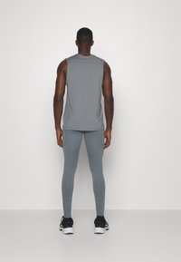 Mann steht mit dem Rücken zur Kamera, trägt ein ärmelloses graues Oberteil, enge graue Leggings, schwarze Socken und schwarz-weiße Laufschuhe vor einem weißen Hintergrund.