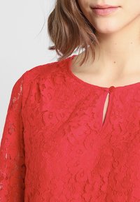 Czerwona koronkowa bluzka z kwiatowymi wzorami, okrągłym dekoltem i małym otworem na guziki z przodu. Miękka w dotyku i o luźnym kroju.