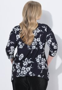 Frau mit langen, welligen blonden Haaren, die ein schwarz-weißes, floral bedrucktes Oberteil und schwarze Hosen trägt, vor einer weißen Wand mit den Händen in die Hüften gestützt.