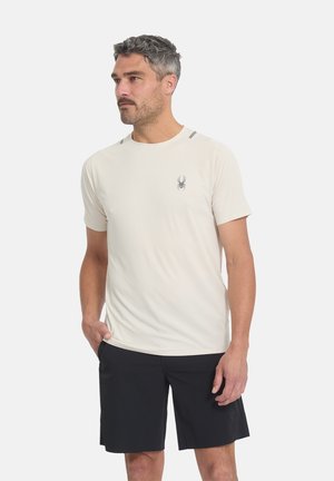 Homme aux cheveux gris courts portant une chemise beige clair à manches courtes avec un logo d'araignée et un short noir jusqu'aux genoux, debout avec une main dans la poche.