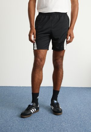 Man draagt zwarte sportshorts met witte zijpanelen, zwarte Adidas-sokken en zwarte Adidas-sneakers op een blauwe tapijtvloer.