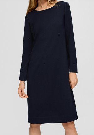Kvinde iført en tekstureret, langærmet, knælængde navyblå kjole med rund hals, stående mod en ensfarvet baggrund.